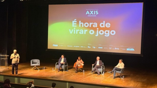Foto que mostra fundo de palco escrito "AXIS &eacute; hora de cirar o jogo" Quatro pessoas sentas em cadeiras *3 homens e uma mulher) e uma em p&aacute;. Todos em cima do palco. 