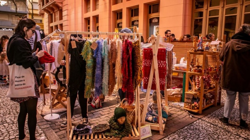 Imagem de uma feira de rua. Nela uma arara om roupas e uma mulher olhando as roupas. Outras pessoas transitando no local.