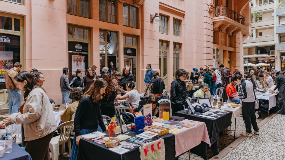 Fotografia retirada na travessa dos cataventos. Na imagem, 7 mesas com produtos de arte gr&aacute;fica como desenhos, pinturas em tecidos, entre outros itens. Em torno das mesas, os vendedores, artistas, e o p&uacute;blico visitante. Em torno de 30 pessoas na foto que tem como fundo, a Casa de Cultura Mario Quintana. 
